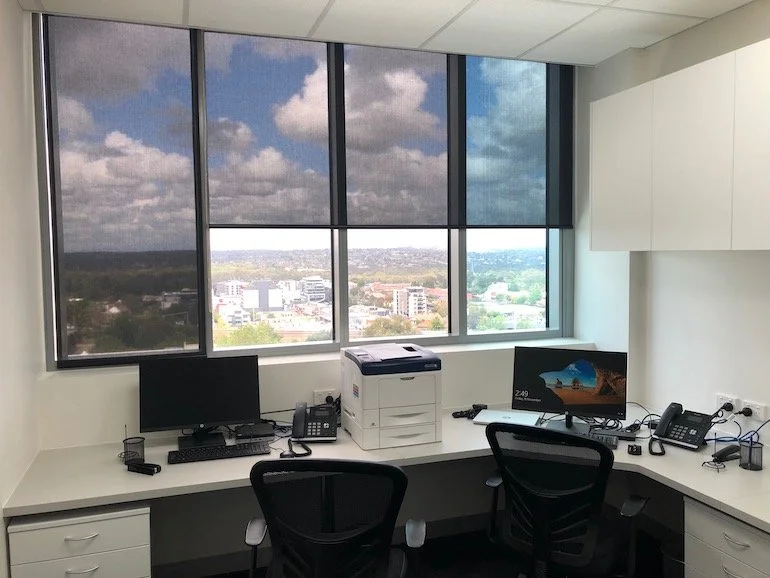 Office cubicle with a large window view of a cityscape, two computer monitors, a printer, phones, and office supplies on the desk.