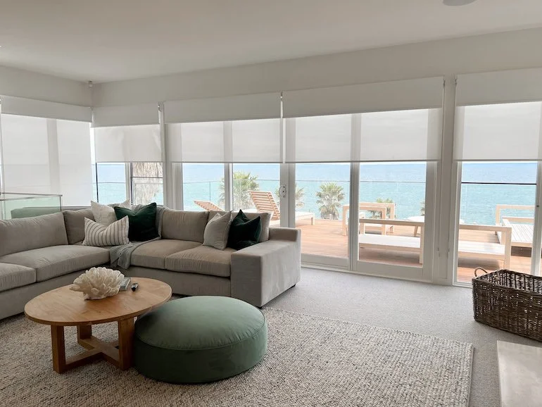 Living room with panoramic ocean view, beige sofa with cushions, wooden coffee table, round green ottoman, balcony with wooden deck chairs, large windows with blinds, and a rattan storage basket.