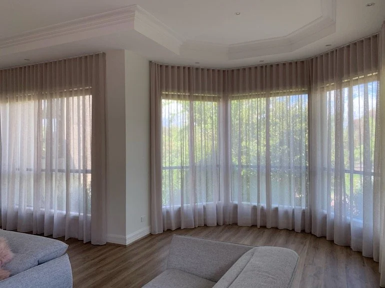 Bright living room with large windows covered by sheer white curtains, hardwood floors, and a light-colored sofa.