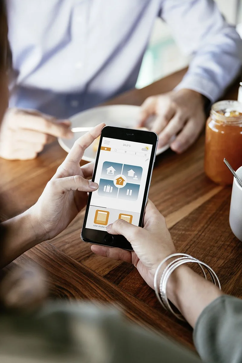 Person holding a smartphone with a home automation app open, showing options for managing home systems, on a wooden dining table with food and a jar of jam.
