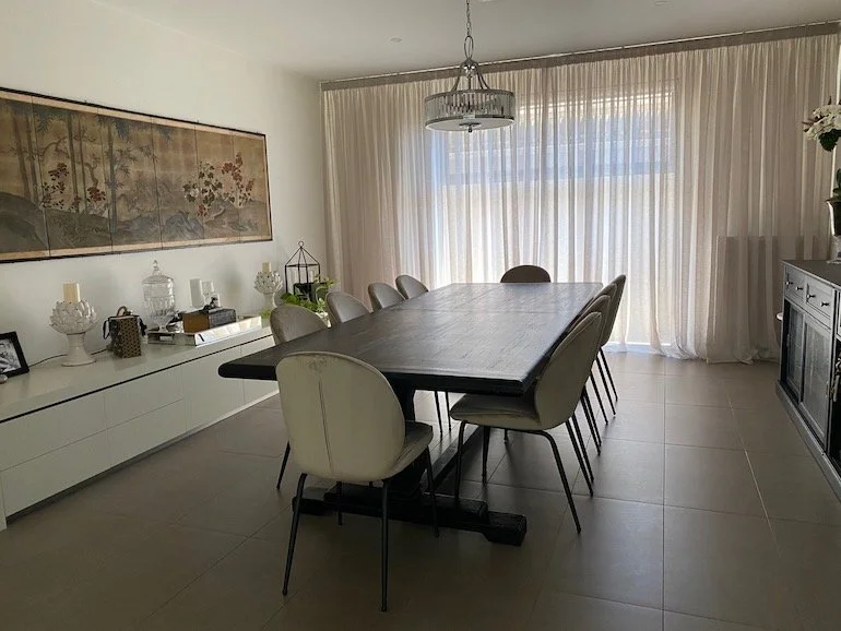 A modern dining room with a long wooden table surrounded by eight beige upholstered chairs, sheer curtains covering large windows, and a white sideboard with decorative items and a large framed artwork on the wall.