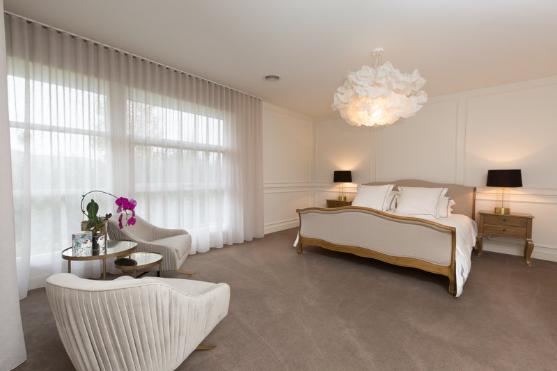 A spacious bedroom with a large bed, two nightstands with black lamps, a sitting area with two white chairs and a small table, and white curtains covering the windows.