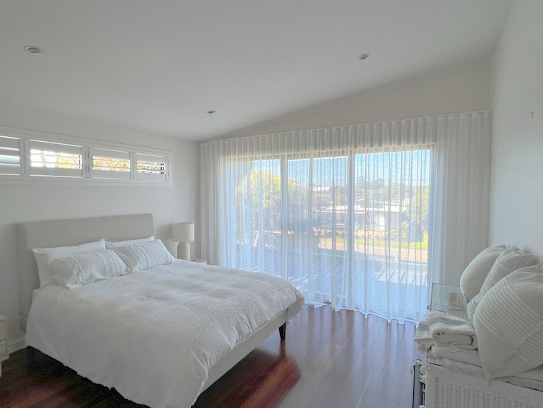 Bright bedroom with white bed, pillows, side table, lamp, and large window with sheer curtains showing outdoor view.