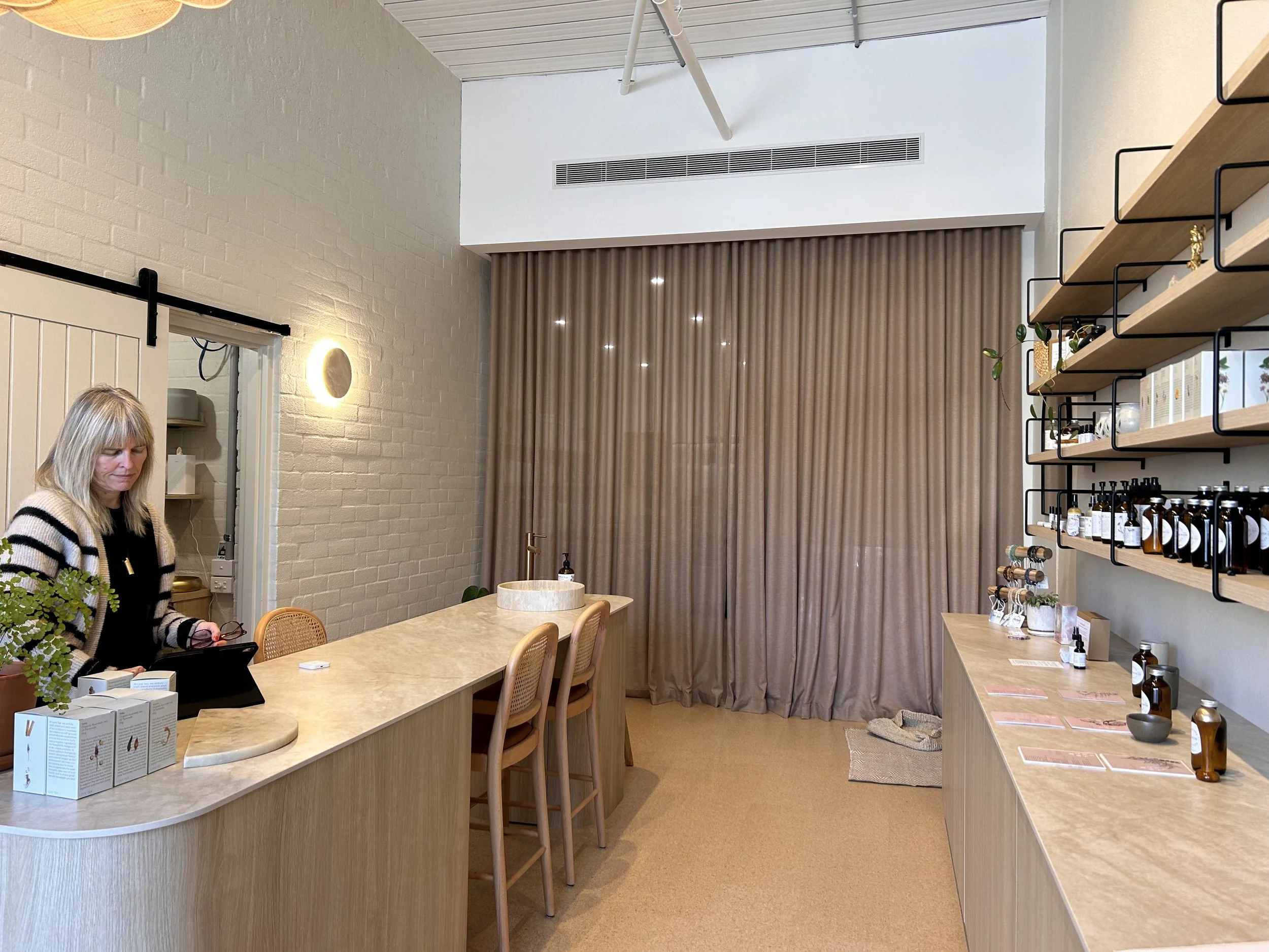 Rasasara founder Sonia Lloyd at the shop counter with full-height clay-coloured curtains and skincare shelves behind her.