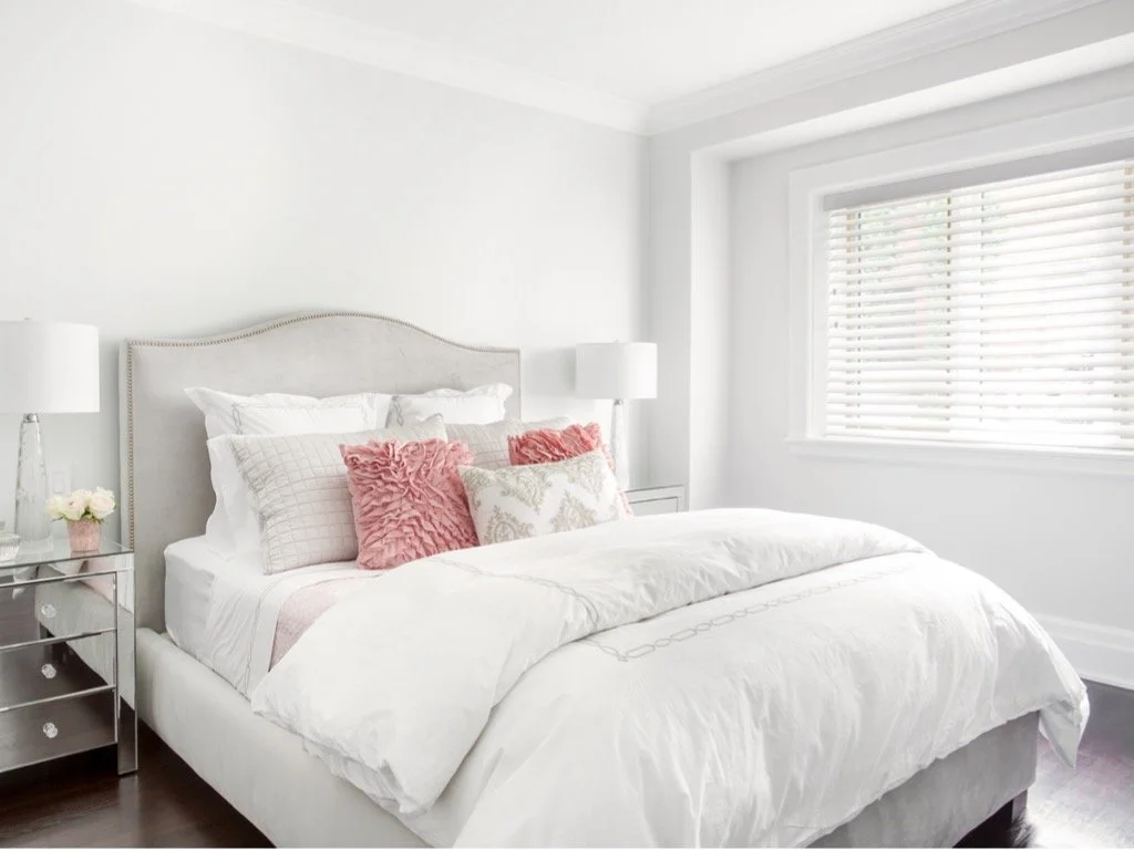 A neatly made bed with white bedding and multiple pillows, two with pink textured covers and one with floral design, in a bright room with white walls and a large window with blinds.