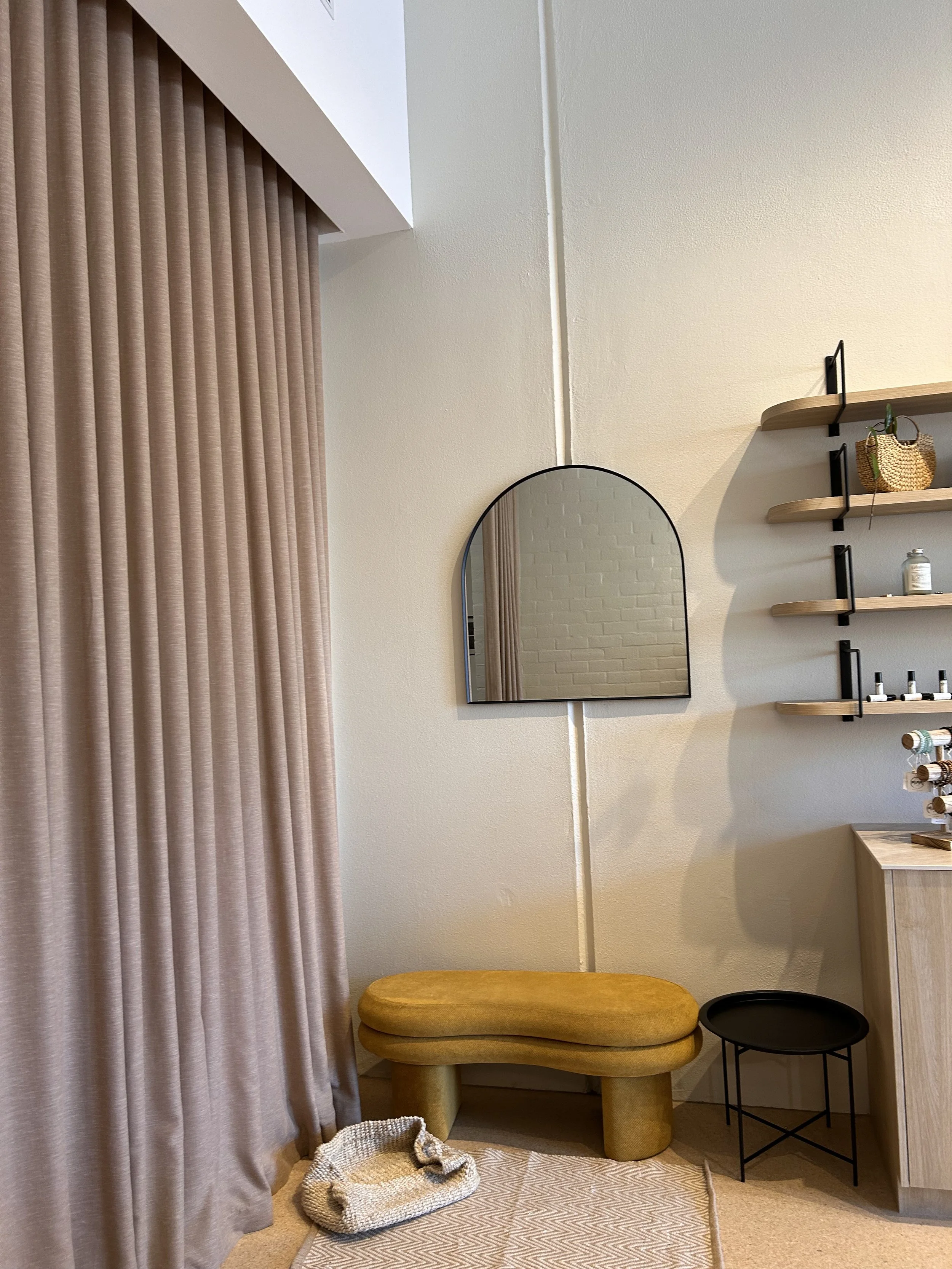Clay-coloured curtains beside an arched mirror, mustard bench and timber display shelves.