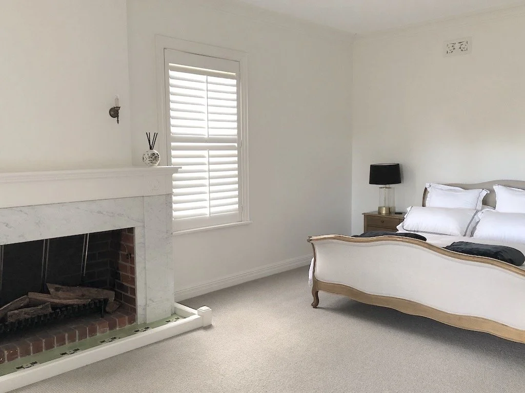 A bedroom with a white bed, black lamp on a wooden nightstand, a window with white shutters, and a white fireplace with a decorative vase and reed diffuser.