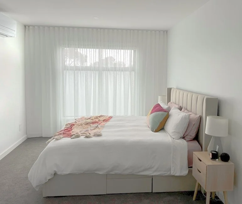 Minimalist bedroom with white walls, large window with sheer curtains, white bed with pink and patterned pillows, beige nightstand, and a white lamp.