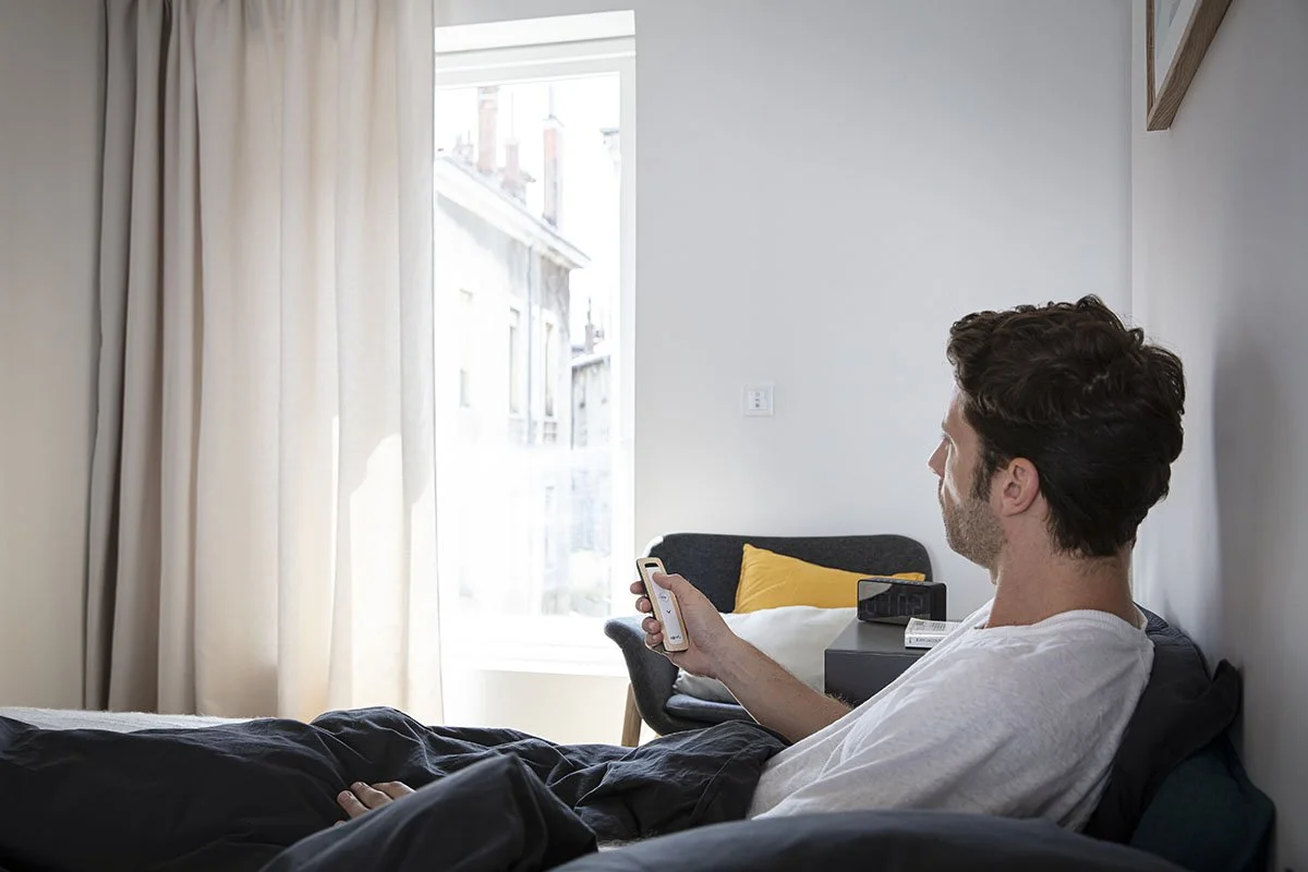 A man with dark curly hair and a white t-shirt lying on a bed, holding a smartphone. The room has a large window with cream-colored curtains, and a side table with a radio, a box, and a yellow pillow on a chair.