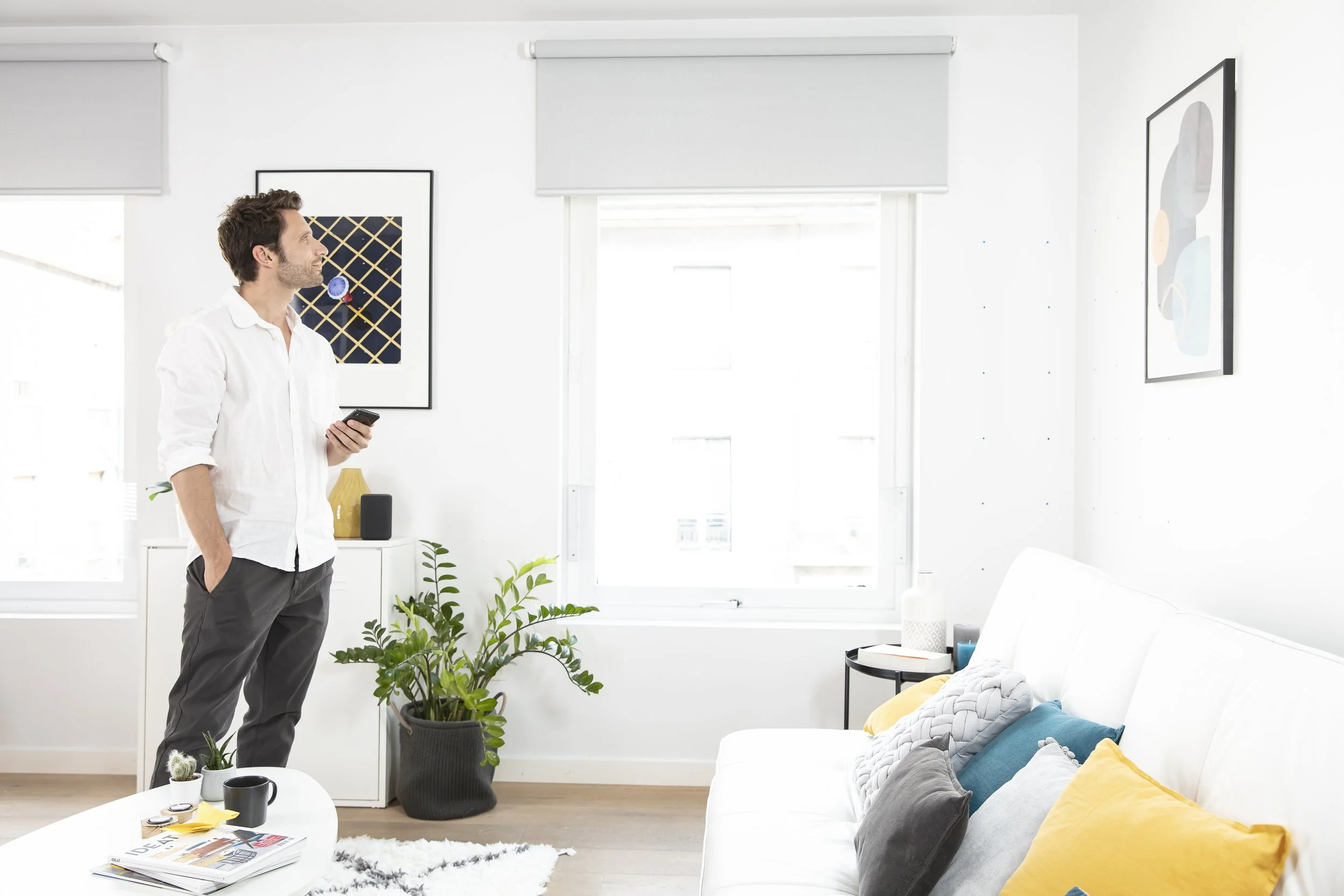 A man standing in a bright, modern living room, holding a smartphone in his right hand, wearing a white shirt and gray pants, with a potted plant and a coffee table in front of him.