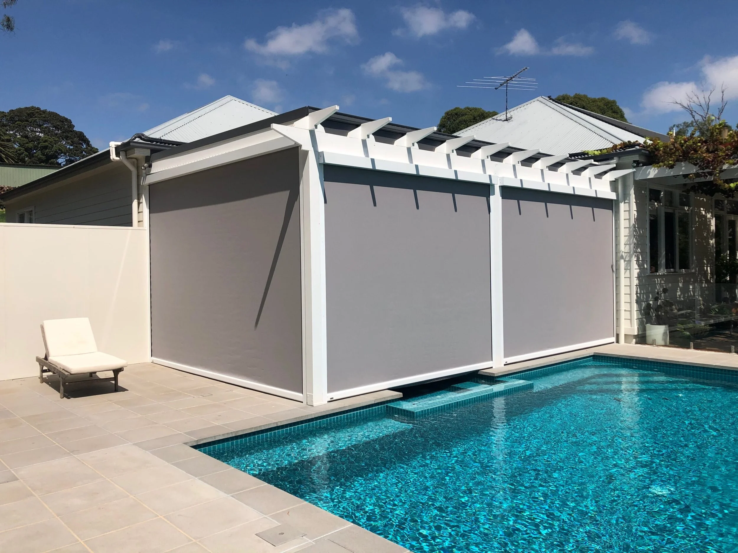 Residential backyard with a swimming pool, gray shade sails, a lounge chair, and a house with white siding and a metal roof.