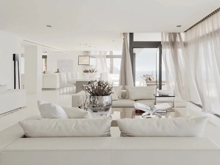 Bright, modern living room with white furniture, sheer curtains, and large windows showing a sea view.