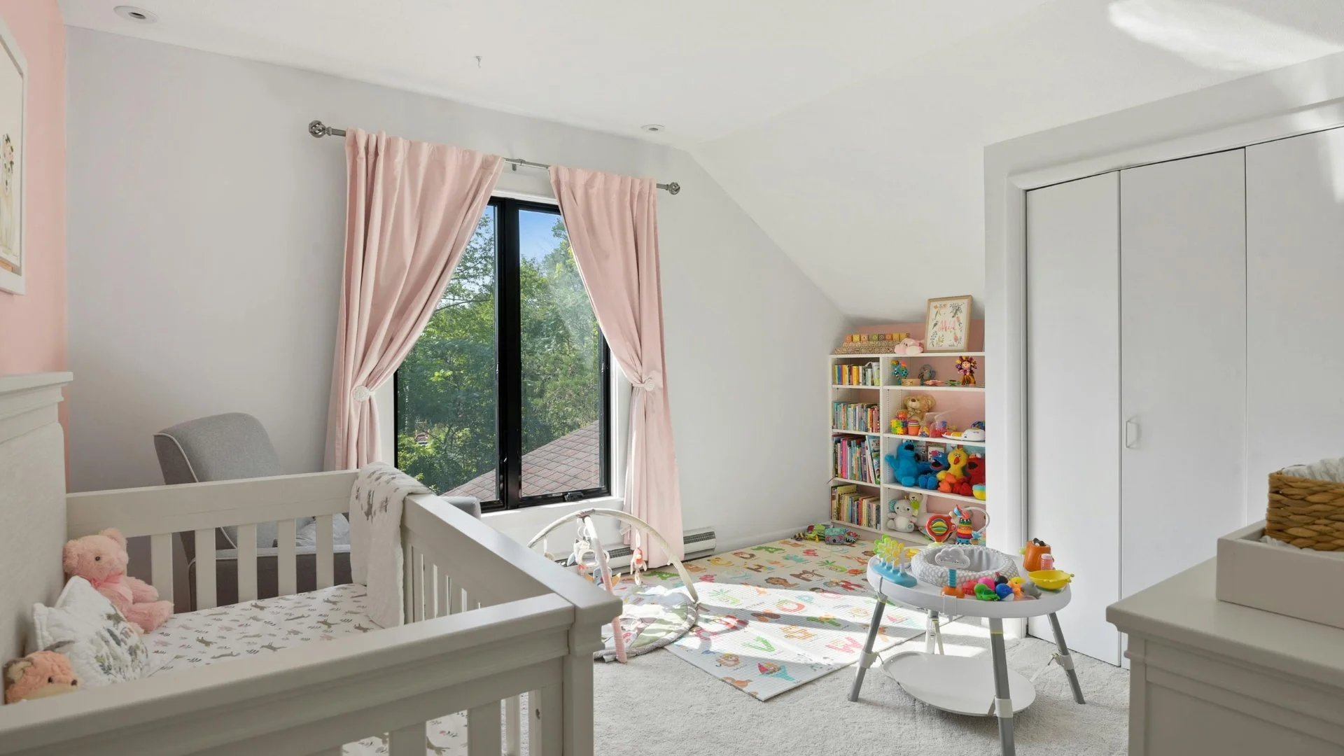Bright children’s bedroom with pale pink curtains, a cot, toys, bookshelves and a colourful play mat.