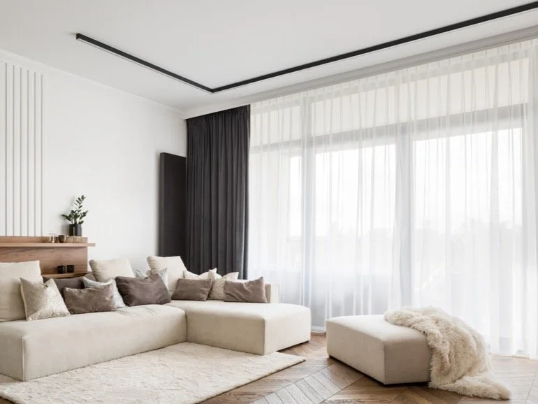 Modern living room with large beige sectional sofa, multiple neutral-colored pillows, sheer white curtains, a black accent wall, and a small potted plant on a wooden shelf.
