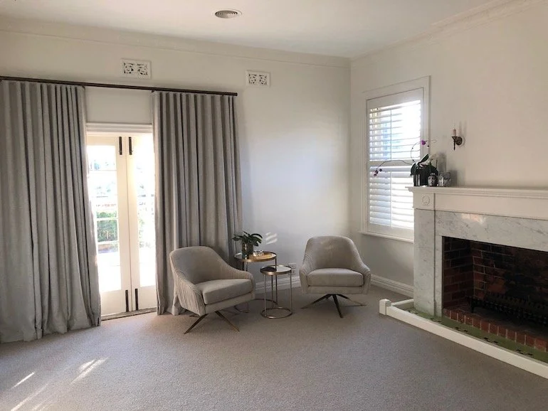 Living room with two light-colored armchairs, a small side table with a plant, a fireplace, and windows with blinds.