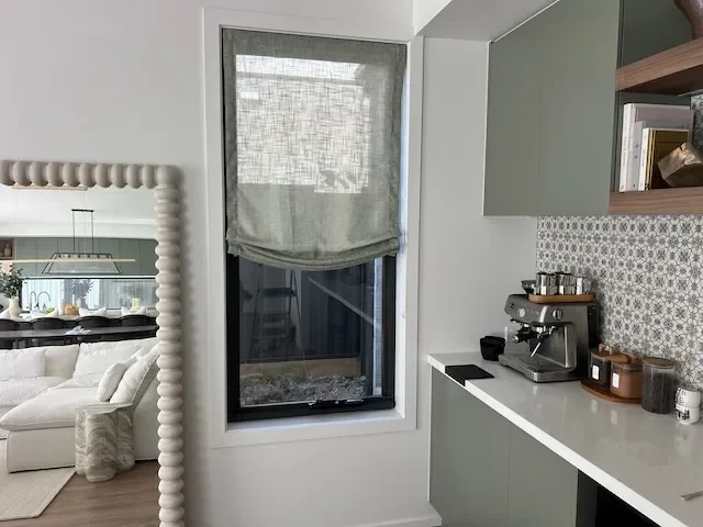 Textured grey Roman blind fitted to a narrow kitchen window beside sage green cabinetry, a white benchtop and coffee station, showcasing a custom window furnishing by Evans Curtains and Blinds in Melbourne.