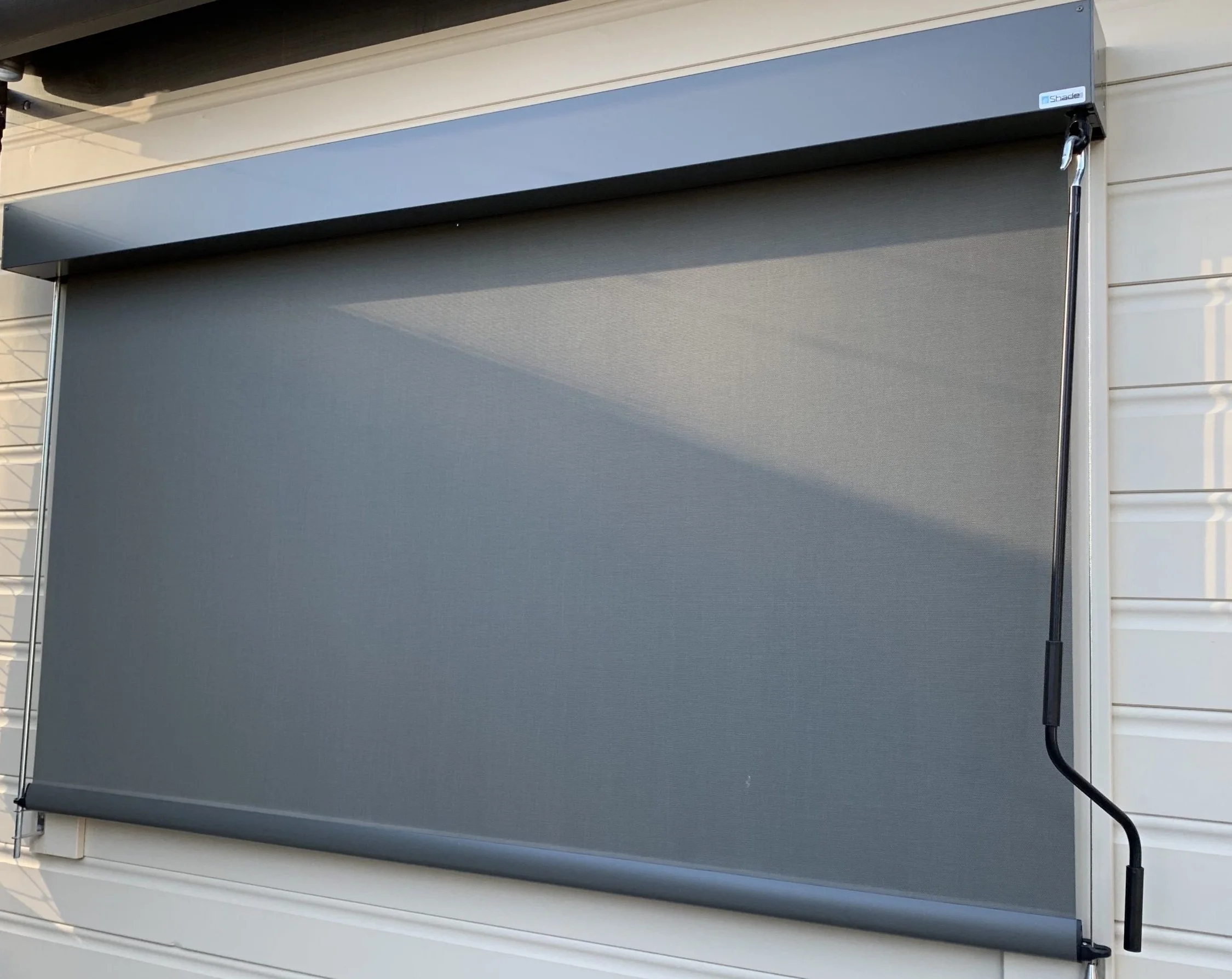 Grey exterior window shade or awning partially rolled down, attached to a white wall.