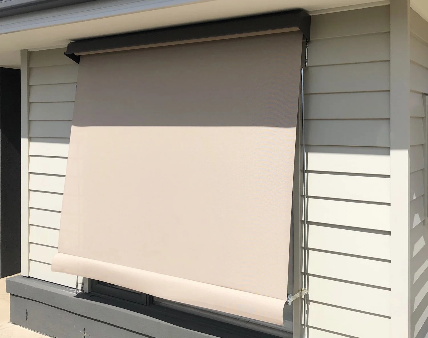 Beige outdoor retractable awning extended over a window or patio area on a house with white horizontal siding.