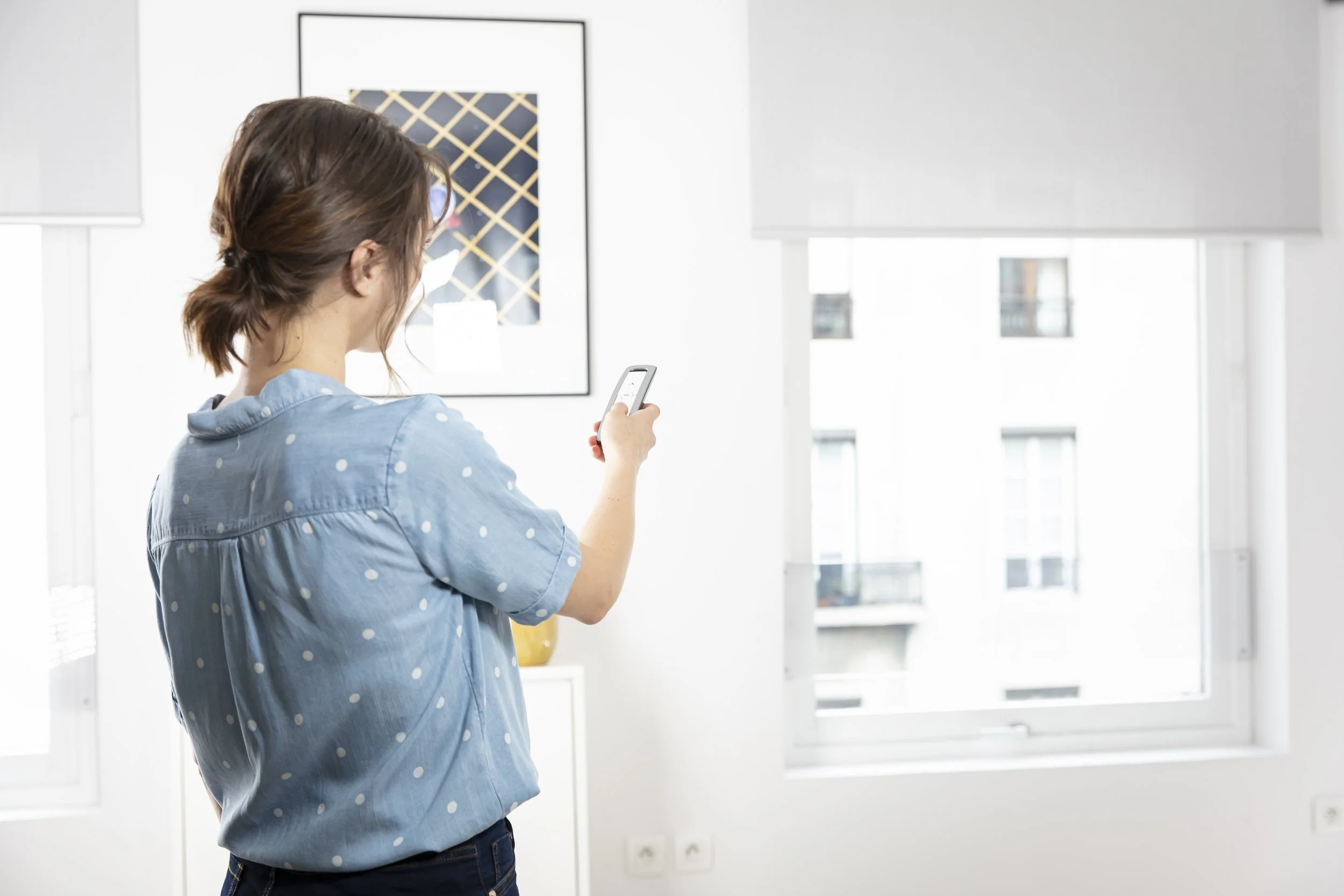 Woman with brown hair tied back, wearing a light blue polka dot shirt, standing indoors and looking at her smartphone.
