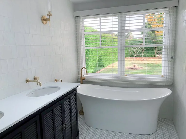 Bathroom with white walls, a bathtub, a large window with blinds showing a green outdoor landscape, a black vanity with a white countertop, and brass fixtures.
