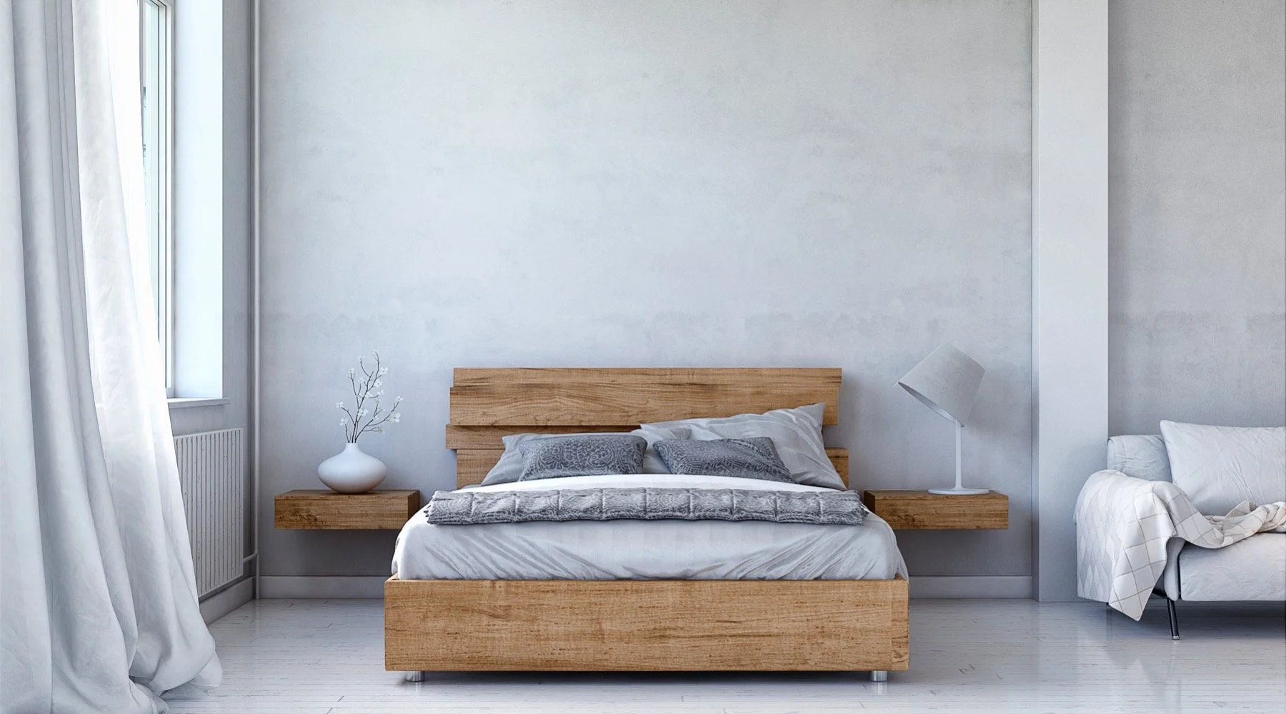 A minimalist bedroom with a wooden bed, gray bedding, two gray pillows, a white vase with branches on the left nightstand, and a white lamp on the right nightstand. A white and beige sofa with a blanket is on the right side, and a window with white curtains is on the left.