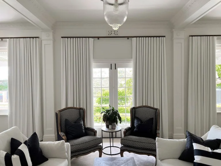 Living room with white walls, large window with white curtains, two armchairs with black pillows, and a small round table with a plant in the center.