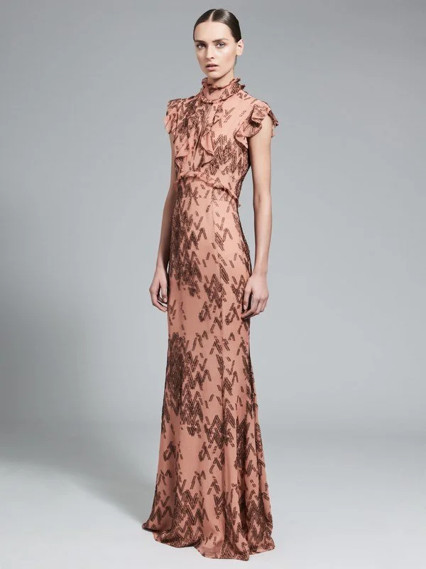 A woman in a long, peach-colored dress with ruffles on the shoulders and a pattern of dark brown chevrons and zigzags stands against a plain gray background.