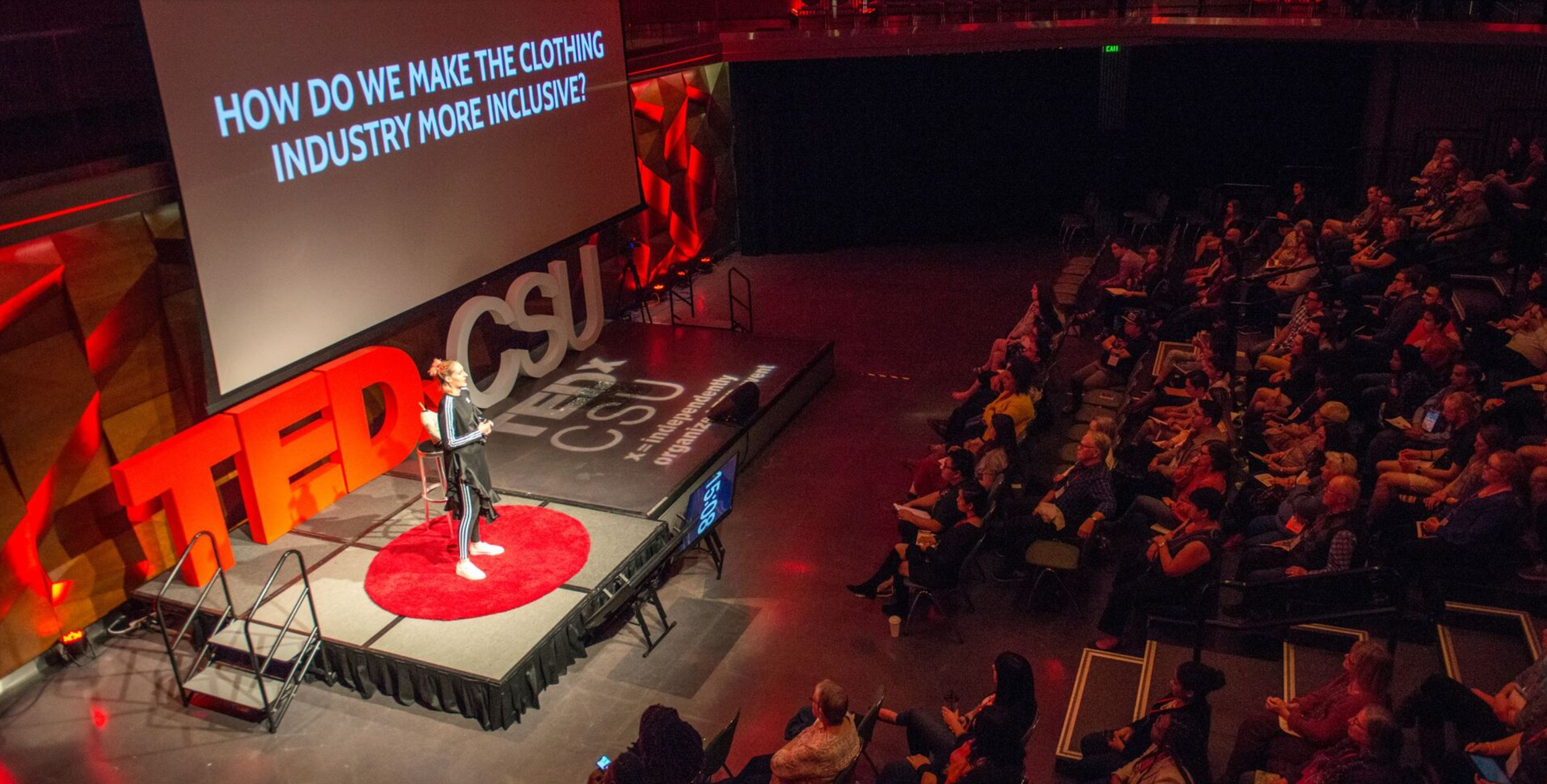 A speaker on a TEDx stage in front of a large screen displays the question, 'How do we make the clothing industry more inclusive?'. The stage features large red TED letters and a dark backdrop. Audience members are seated facing the stage, some taking notes or using laptops.
