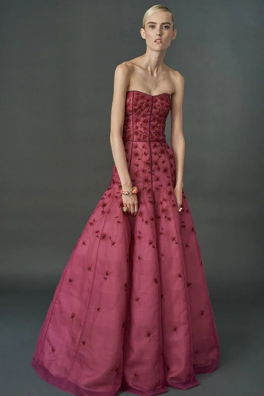 A woman with blonde hair and fair skin wearing a strapless, pink, embroidered ball gown with floral patterns and a full skirt, standing against a gray background.