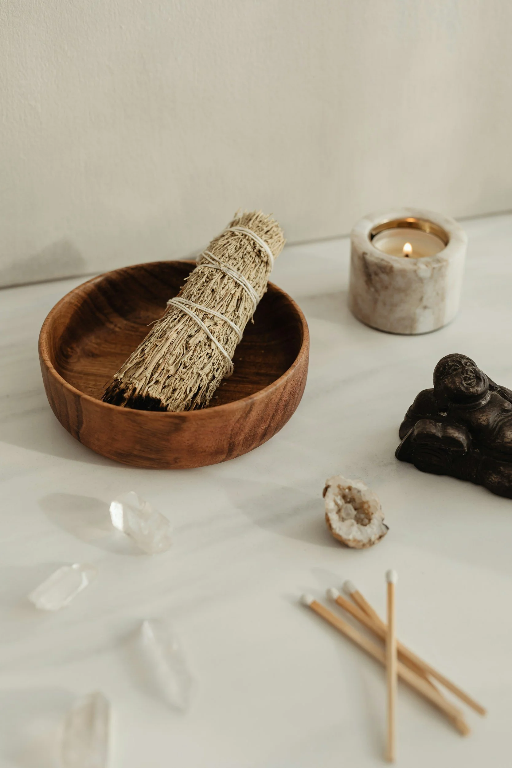 A wooden bowl with a bundle of sage, a lit candle in a marble holder, a small black figure, clear quartz stones, a geode slice, and a bundle of matchsticks on a white surface against a neutral background.
