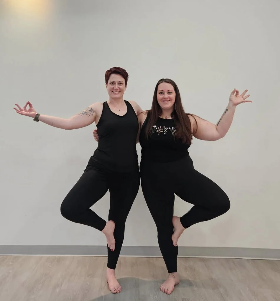 Two women practicing yoga indoors, standing on one leg with the other foot resting on the inner thigh of the standing leg, in a tree pose, smiling at the camera.