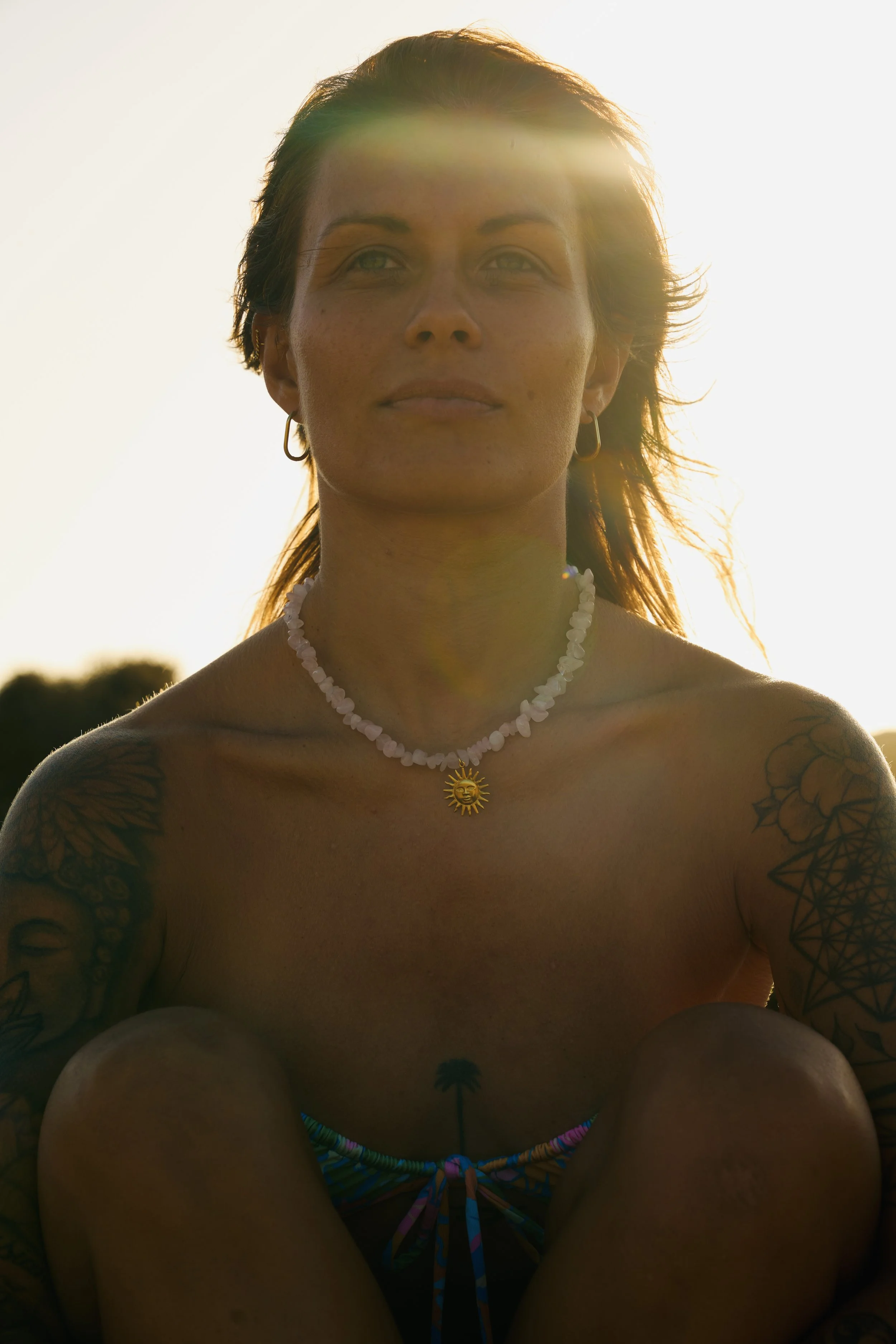 Portrait of a woman with tattoos, wearing a pearl necklace with a sun pendant, in intense sunlight in the background.