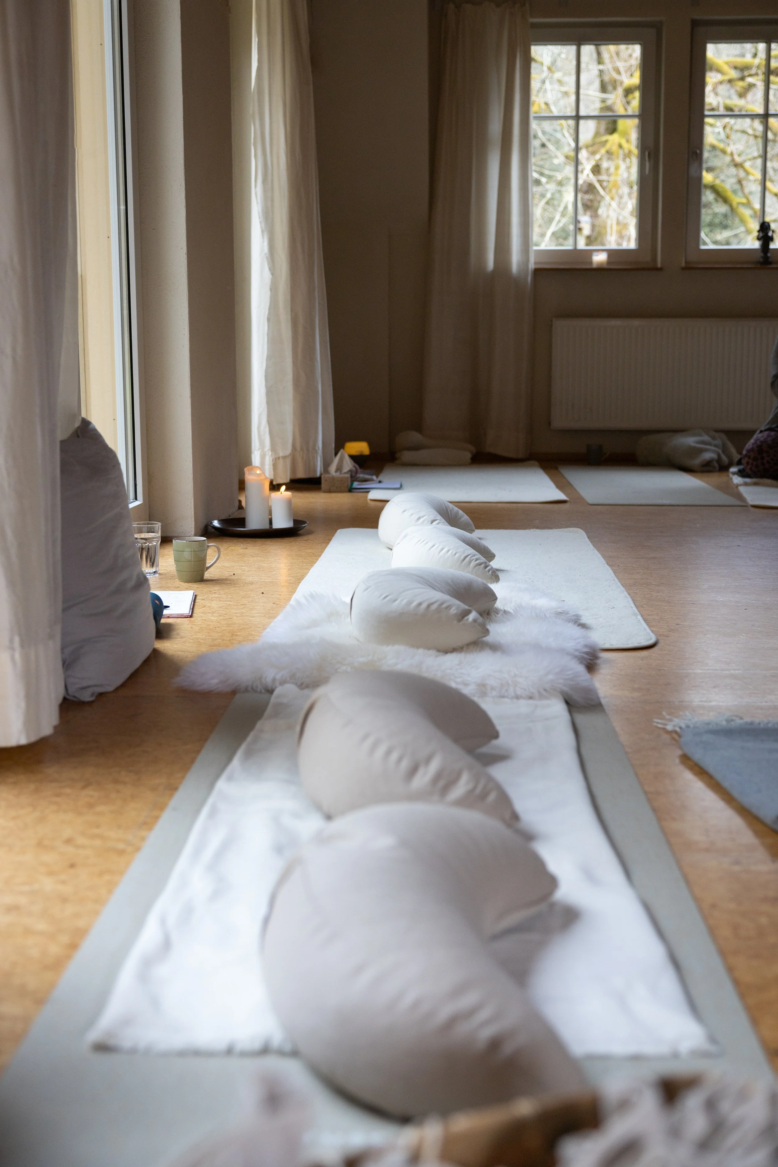 Ein gemütlicher Raum mit weißen Kissen auf einer weißen Decke, Kerzen, einer Tasse, einem Glas Wasser, und einem Fenster mit Blick auf Bäume.