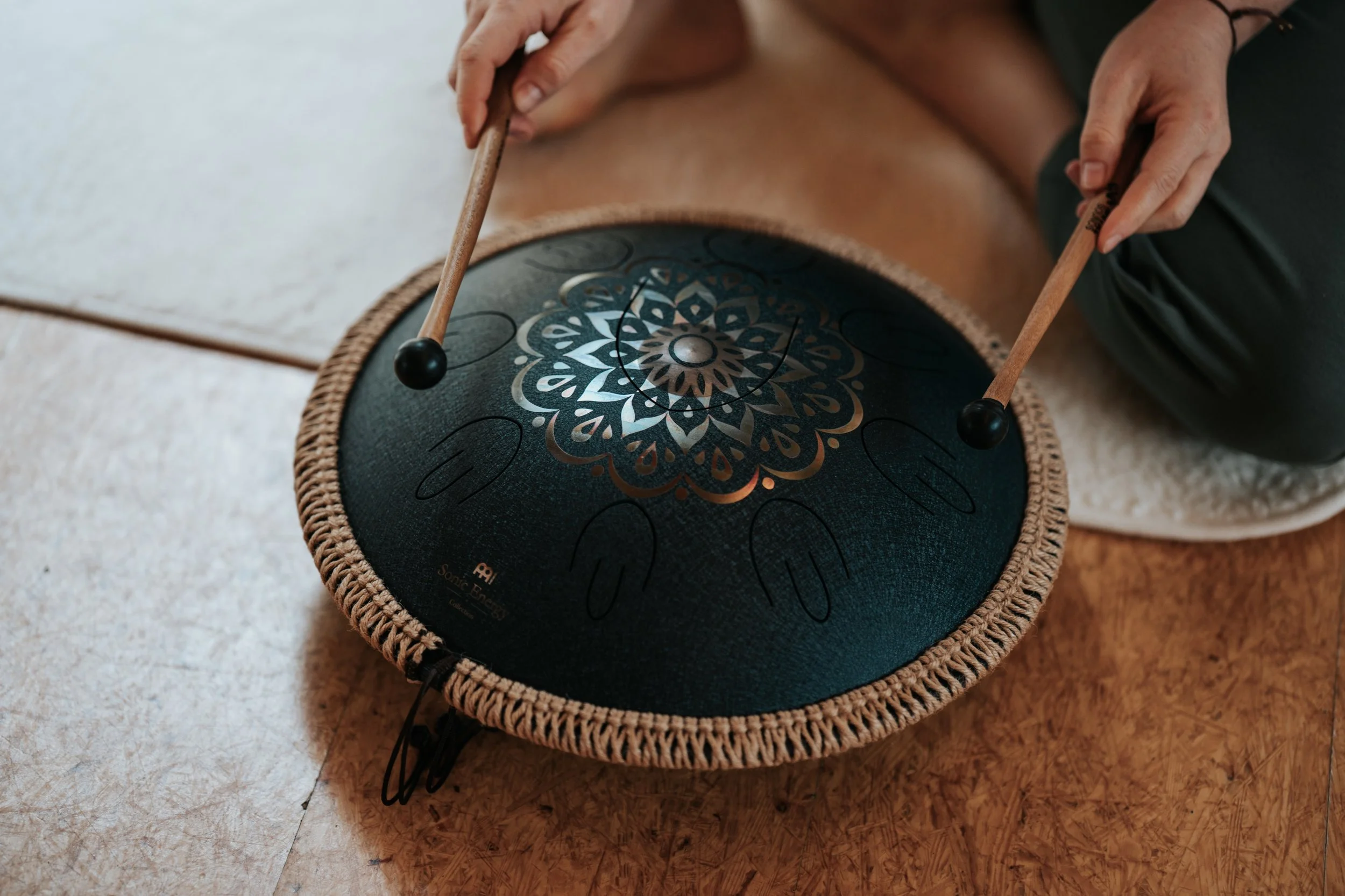 Person spielt auf einer schwarzen Handpan mit goldener Musterung, sitzend auf einem Holzboden.