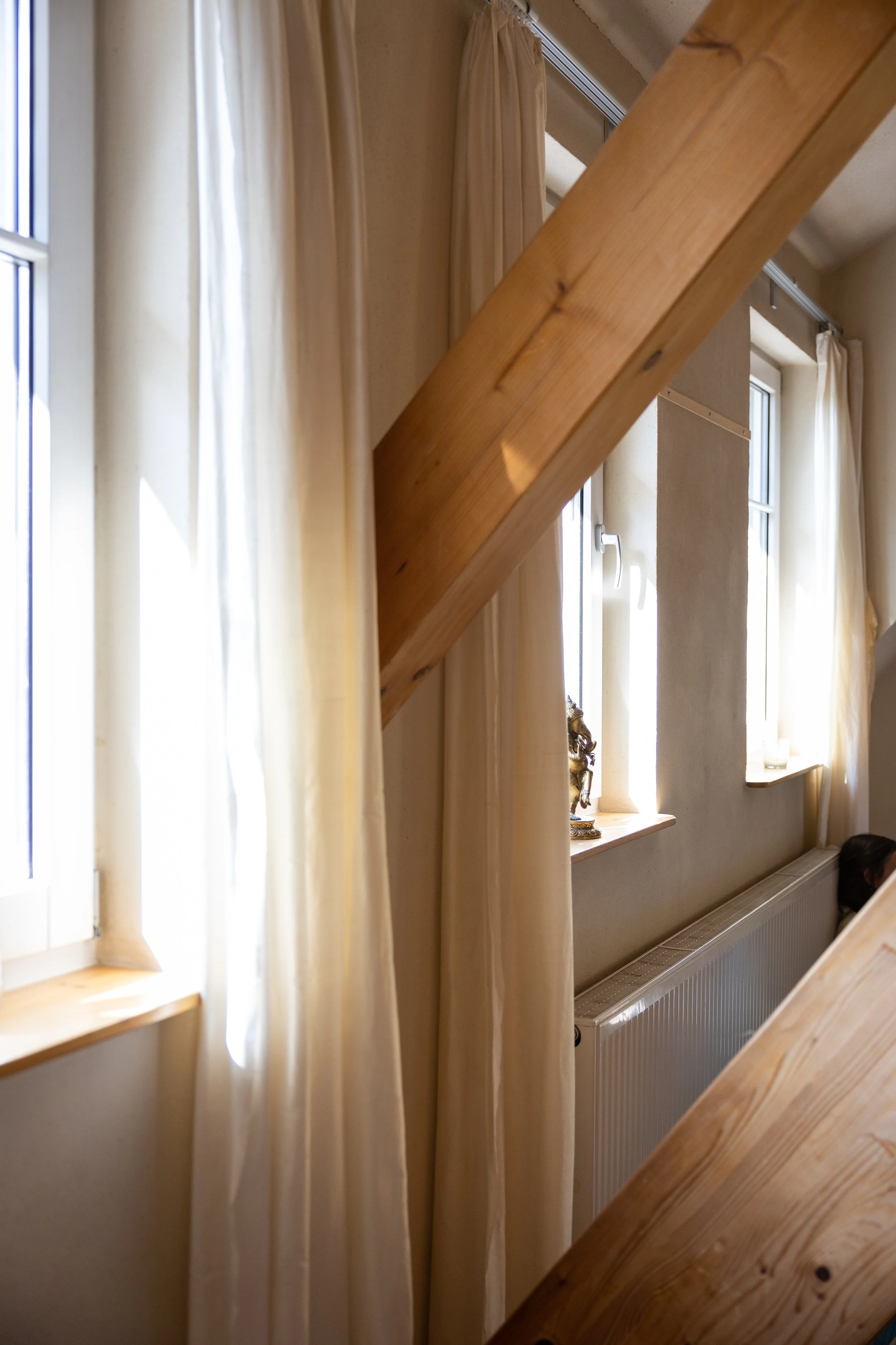 Innenraum mit Fenster, weißen Vorhängen, Holzlampe, Heizkörper und einer Holzstrebe.