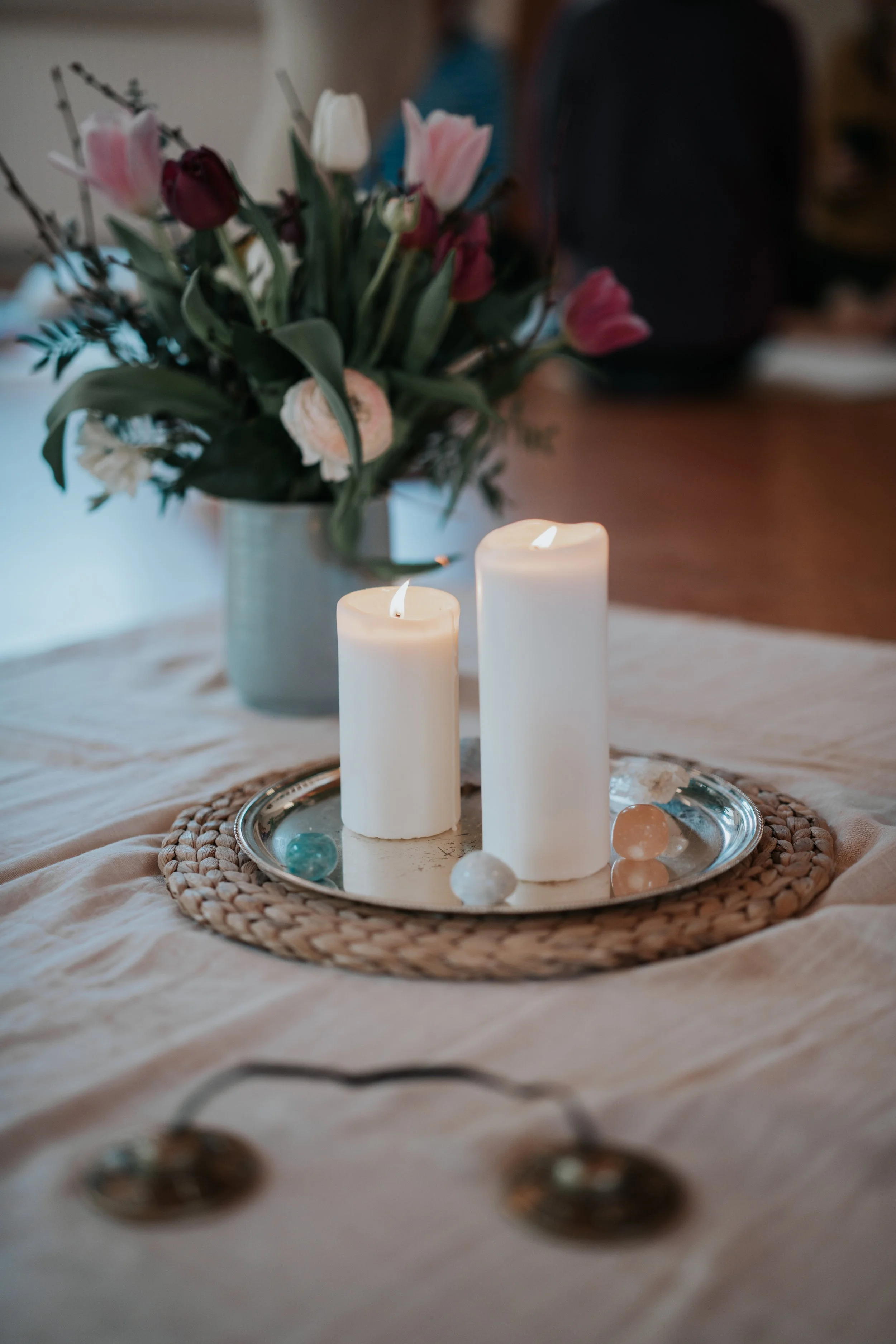 Eine Tafel mit zwei brennenden Kerzen auf einer runden silbernen Schale, umgeben von Kristallen, auf einem Tisch mit einer cremefarbenen Tischdecke. Im Hintergrund eine Vase mit bunten Tulpen.