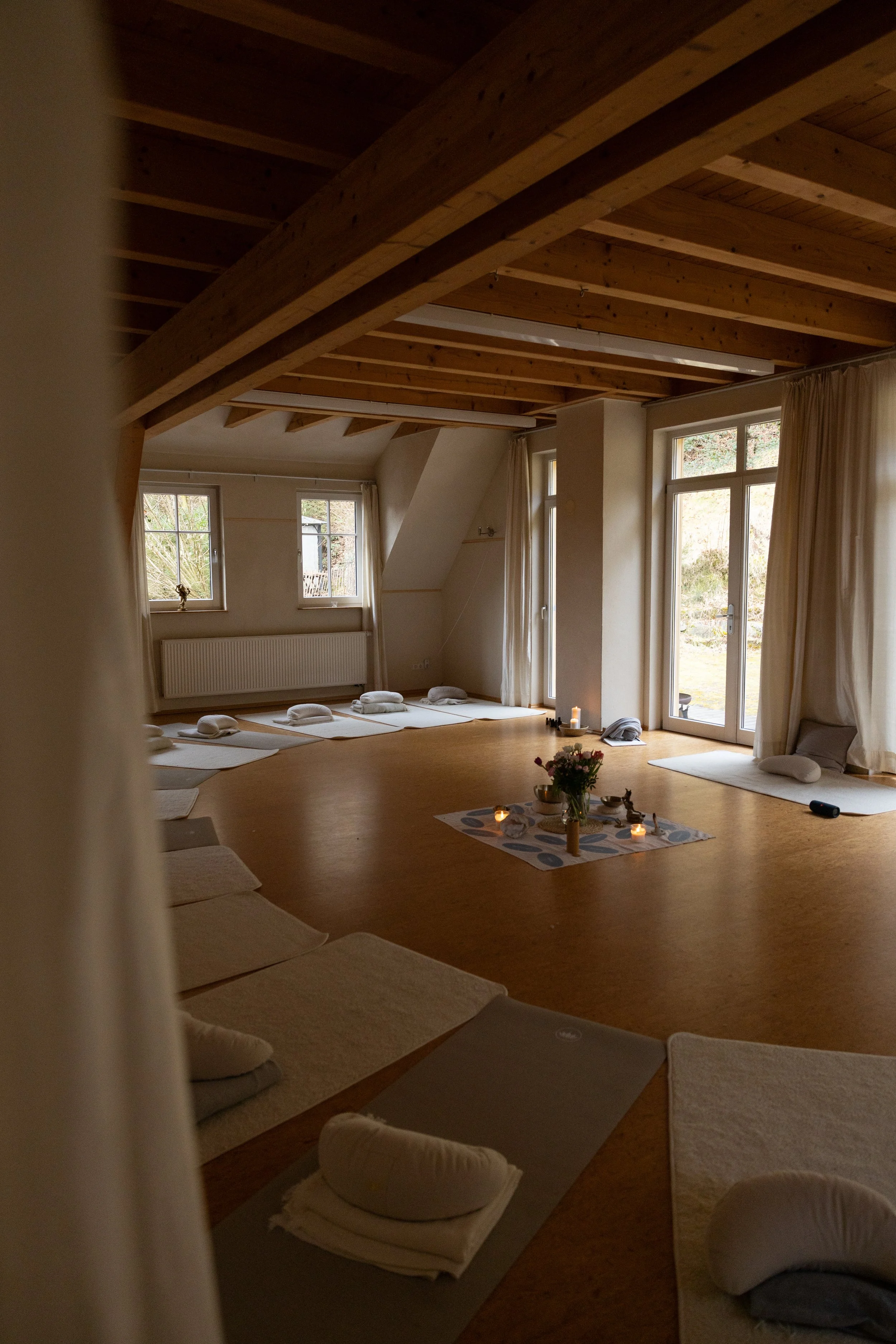 Ein hell erleuchteter Raum mit Holzdecken und Dielenboden, mehrere auf dem Boden liegende Yogamatten mit Kissen, eine kleine runde Ablage in der Mitte mit Kerzen, Blumen und weiteren Gegenständen, große Fenster und eine Tür lassen natürliches Licht h