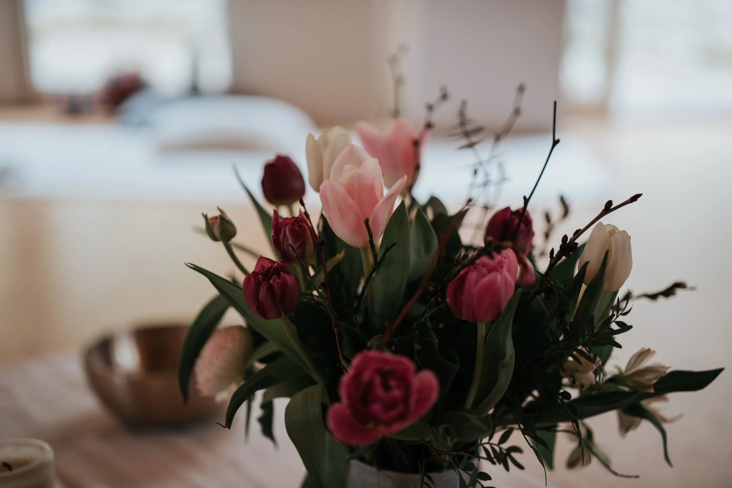 Blumenstrauß mit rosa und weißen Tulpen in einer Vase auf einem Tisch, unscharfer Hintergrund mit Bett und Fenster.