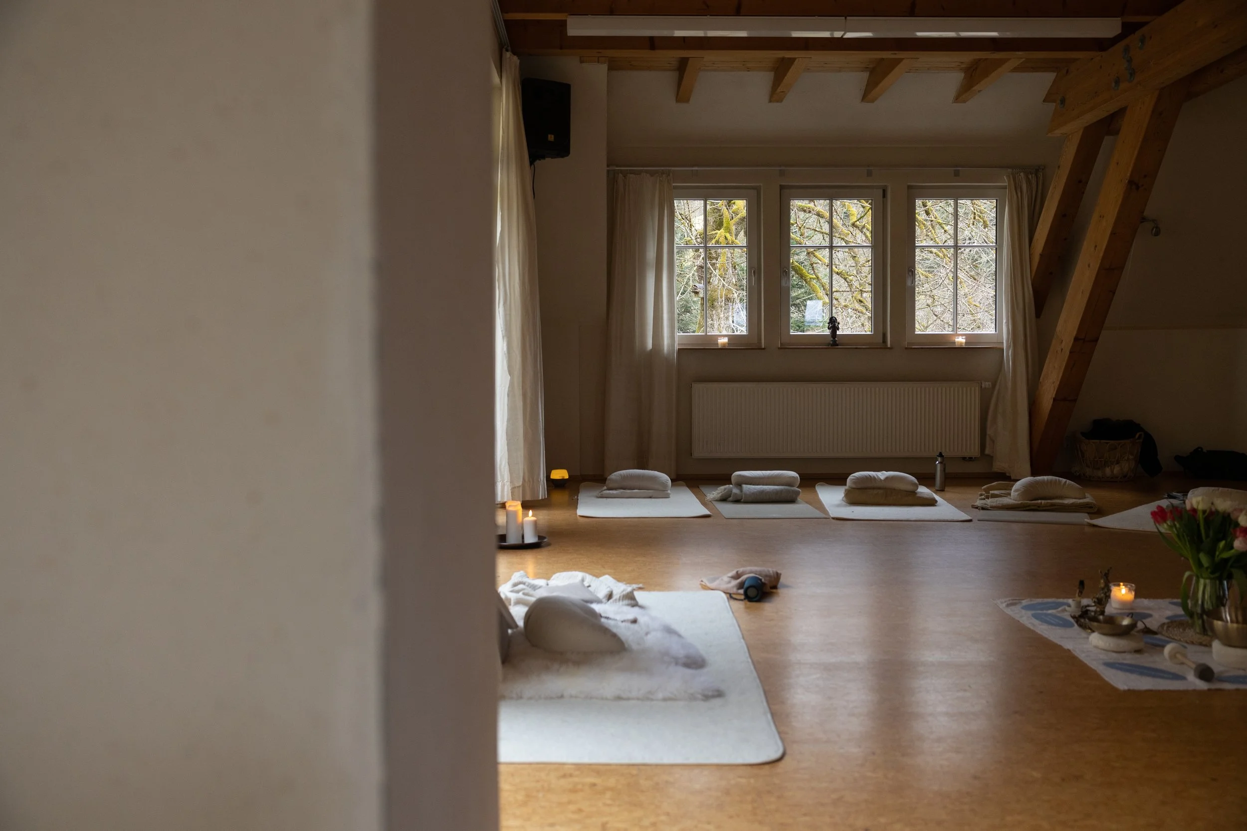 Ein Raum für Yoga oder Meditation mit Matten, Kissen und Kerzen, natürliche Beleuchtung durch Fenster, Holzdecke und -balken, ruhige Atmosphäre.
