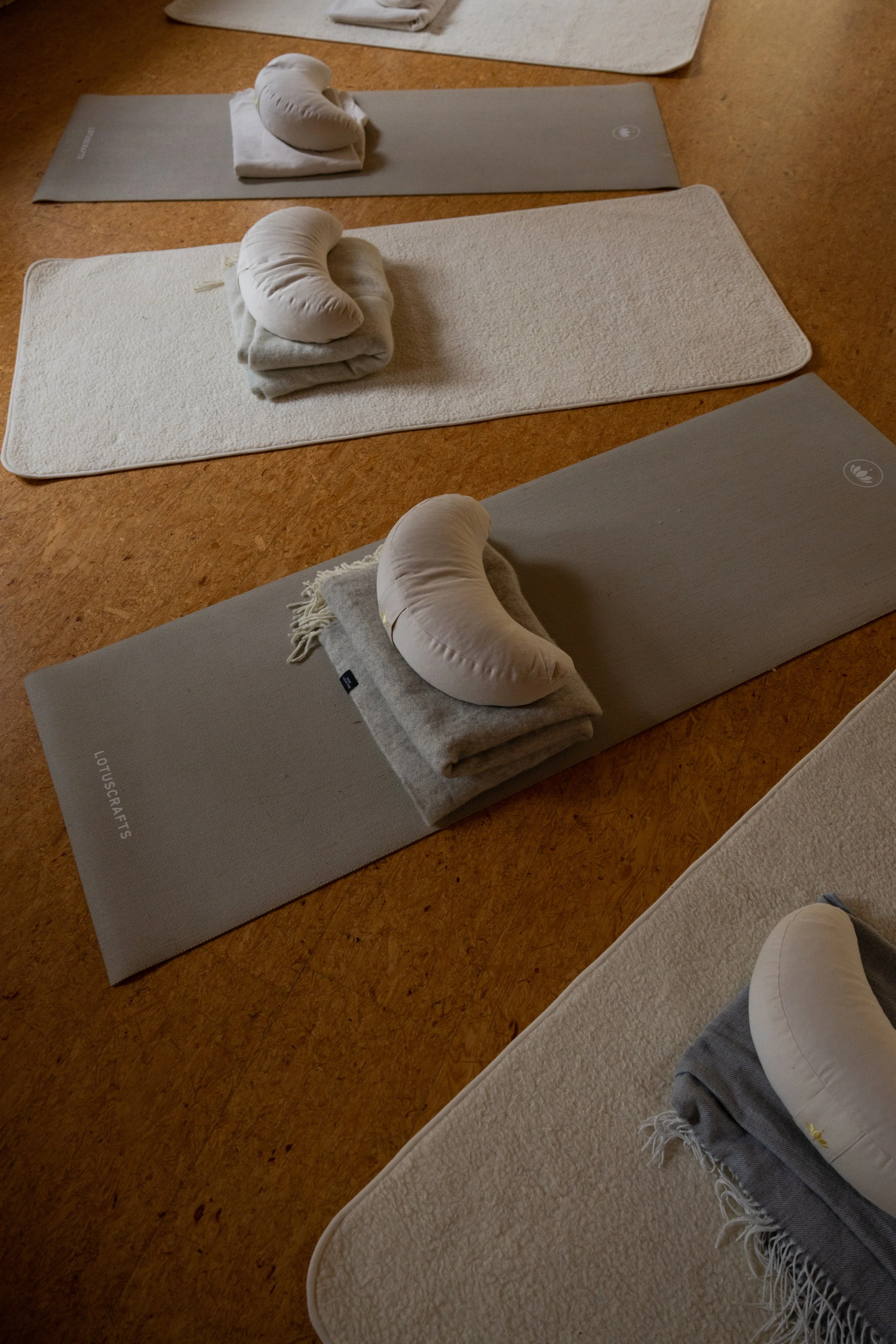 Yogamatten mit zusammengerollten Kissen und Handtüchern, vorbereitet für eine Yoga- oder Entspannungsstunde, auf Holzboden verteilt.