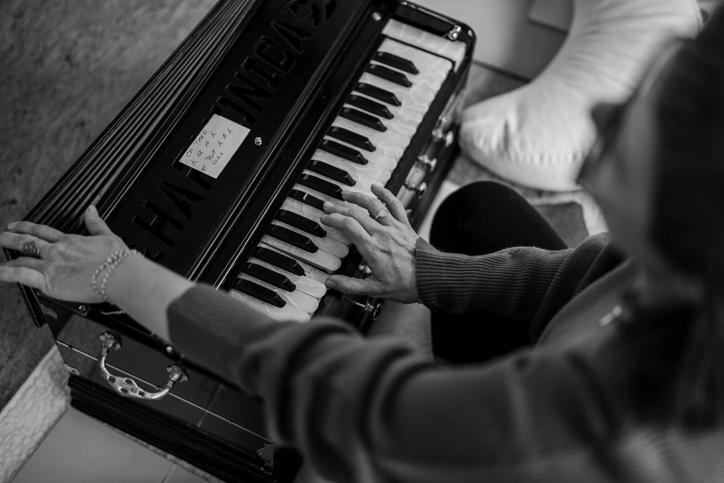 Eine Person spielt auf einem kleinen Klavier mit schwarzer Oberfläche, weißen und schwarzen Tasten. Die Person trägt eine Armbanduhr und ein Armband. Es liegt ein Notizzettel mit handgeschriebenen Notizen auf dem Klavier. Im Hintergrund ist eine Fußs