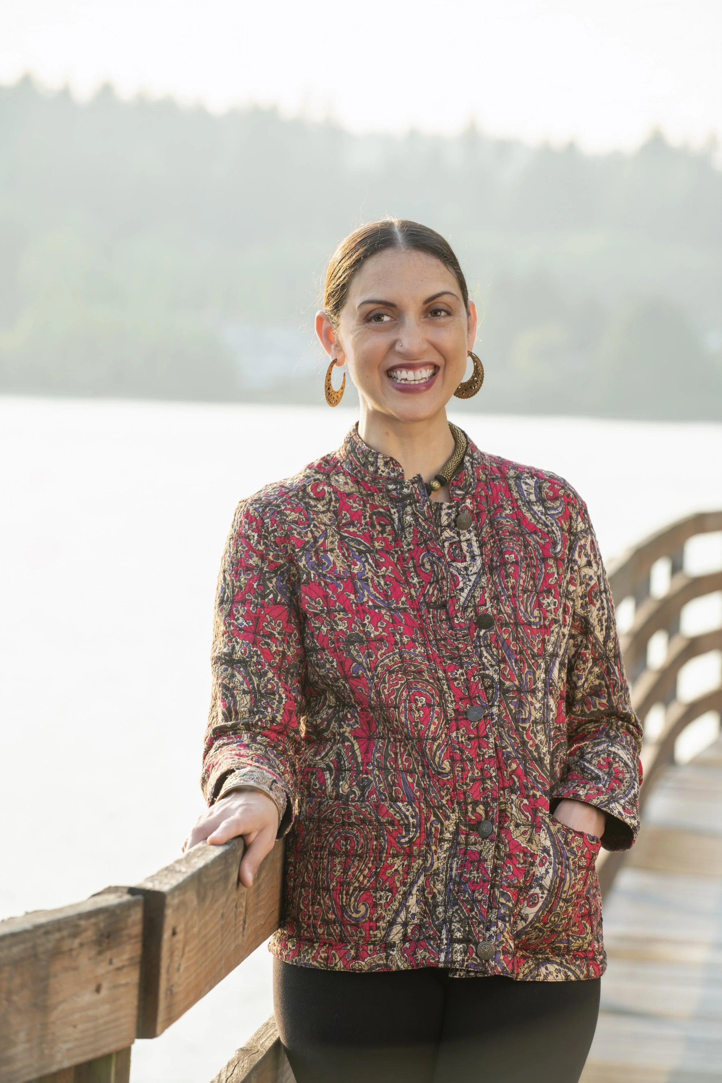 A woman with dark hair tied back, smiling, stands on a wooden dock by a body of water with trees in the background, wearing a colorful paisley-patterned jacket and large earrings.