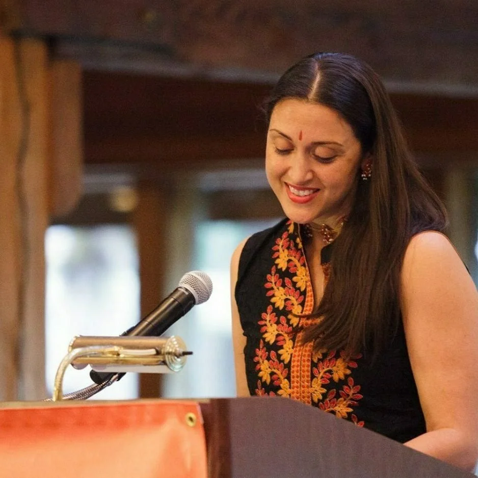 A woman with long dark hair smiling at a microphone, wearing traditional Indian attire with embroidery, indoors in a warmly lit setting.