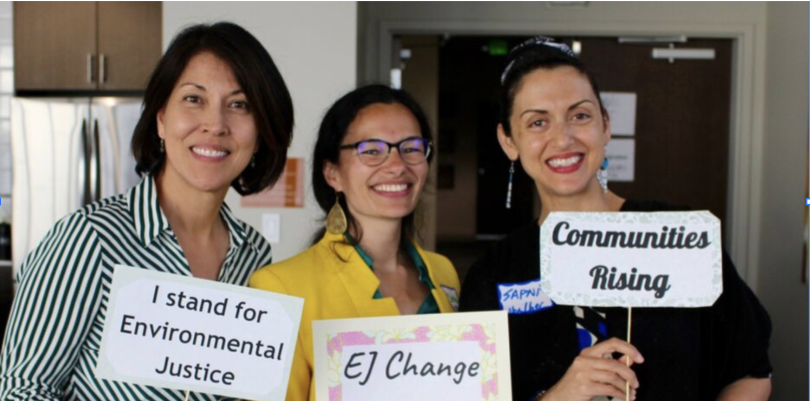 Three women holding signs at a gathering that advocate for environmental justice and community activism. The woman on the left holds a sign that says 'I stand for Environmental Justice,' the woman in the middle's sign reads 'EJ Change,' and the woman on the right's sign says 'Communities Rising.'