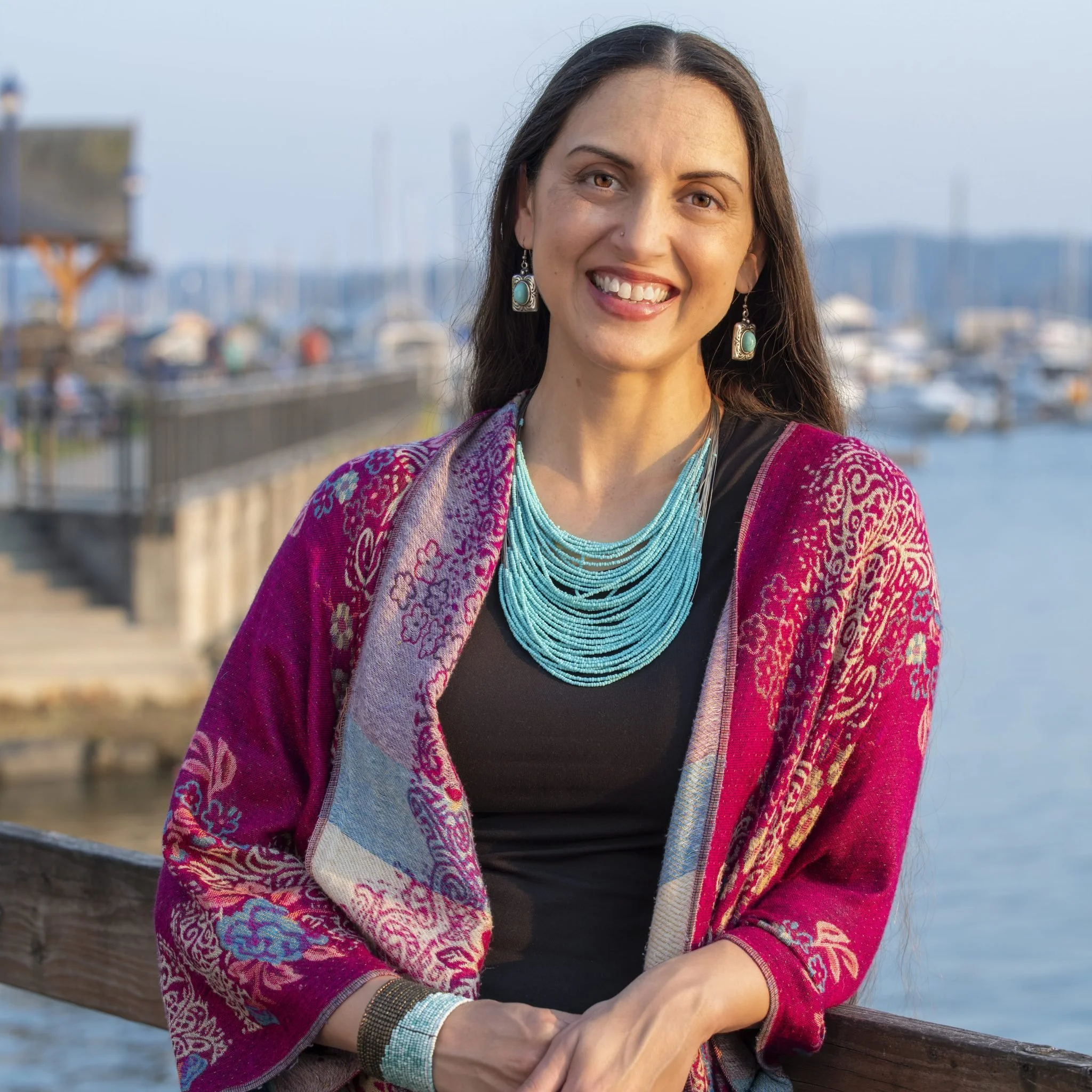 A woman with long brown hair and tan skin smiling outdoors near a body of water, wearing turquoise jewelry and a colorful patterned shawl.