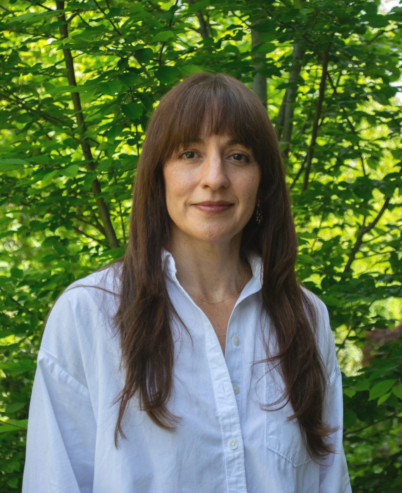 A woman with long brown hair and a white button-up shirt standing outdoors in front of green leafy trees.