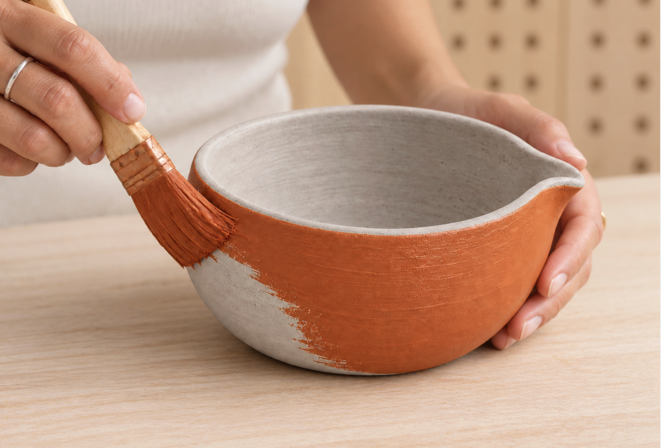 Hände tragen mit einem Pinsel orange Engobe auf die Außenseite eines Matcha-Chawans mit Ausguss auf. Innen bleibt der Ton naturfarben.