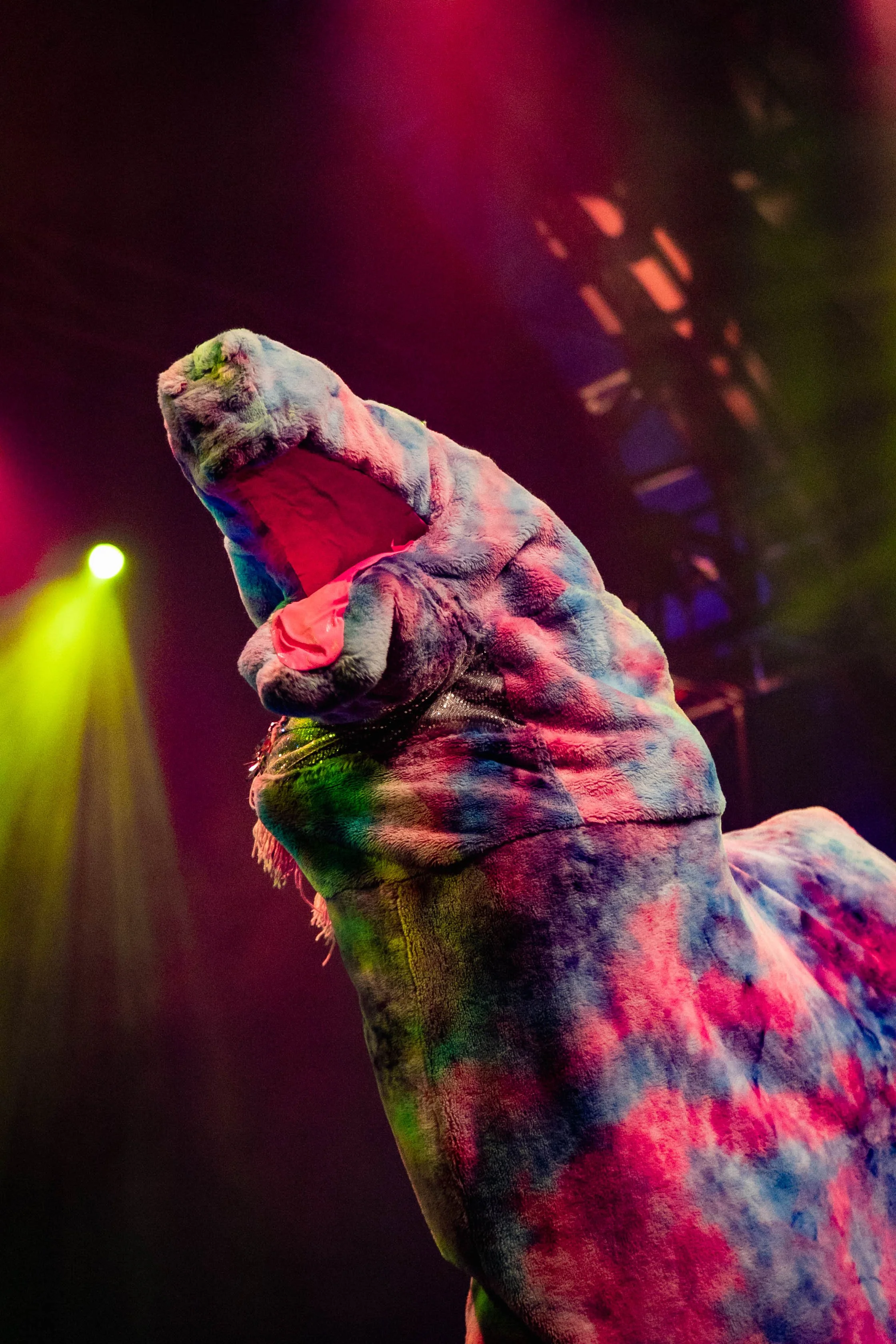 A person dressed in a colorful dinosaur costume performing on stage with multicolored lights.