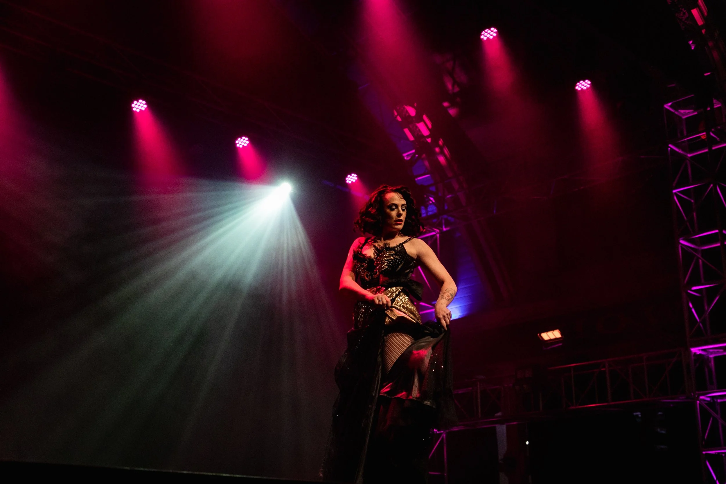 A woman with dark curly hair performing on stage, dressed in a black and gold outfit with fishnet stockings, under stage lighting with pink, purple, and white spotlights.
