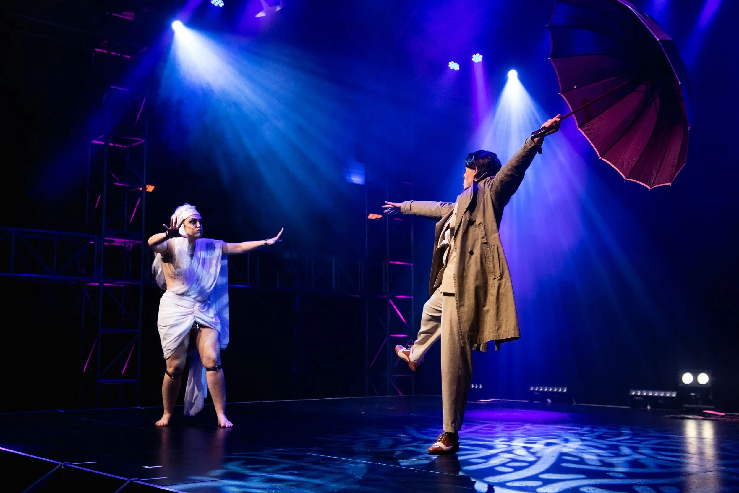 Two performers on stage under blue and purple lights, with one holding a purple umbrella. One performer is dressed in beige with arms outstretched, and the other is in white attire with a white headscarf, reaching out with hands.